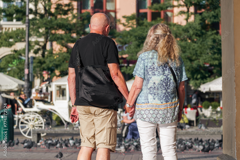 In a busy urban setting, an elderly couple walks hand in hand, a testament to everlasting love and partnership, against the backdrop of a white carriage and doves In a busy urban setting, an elderly couple walks hand in hand, a testament to everlasting love and partnership, against the backdrop of a white carriage and doves
