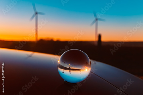 Billede på lærred Crystal ball sunset with wind power plant silhouettes and reflections on a car r