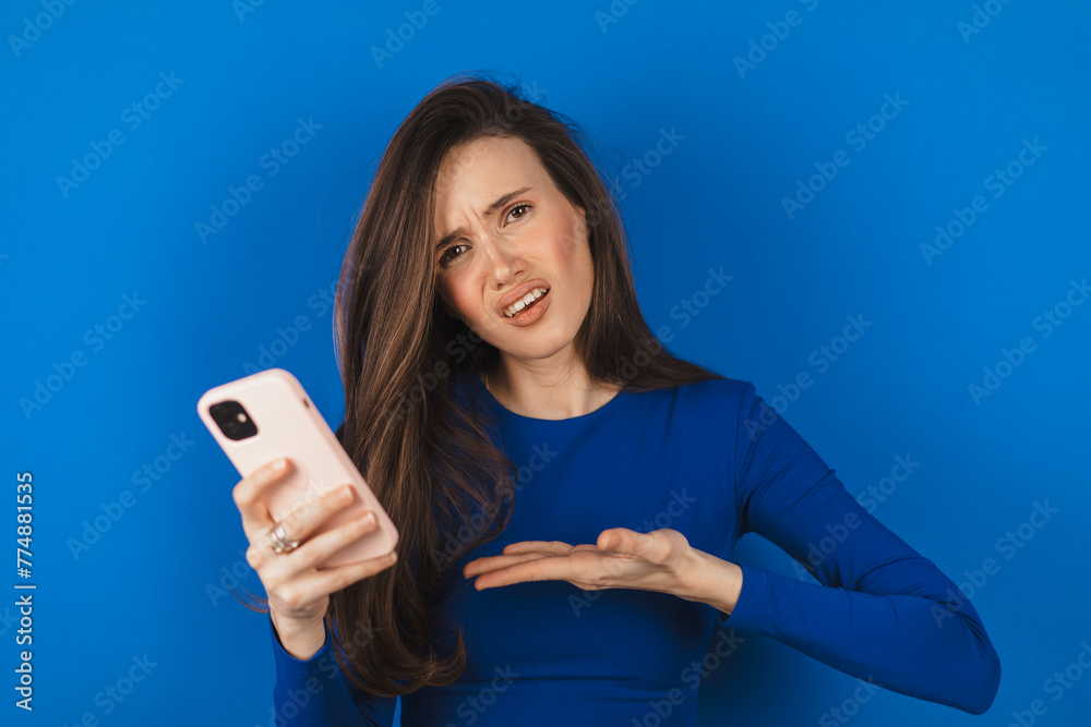 Mad and shock young brunette beautiful woman using mobile phone isolated on blue background. Yelling unhappy woman hold mobile phone. Disappointed sad upset lady horrified impressed news. WTF. Oh no. Mad and shock young brunette beautiful woman using mobile phone isolated on blue background. Yelling unhappy woman hold mobile phone. Disappointed sad upset lady horrified impressed news. WTF. Oh no.