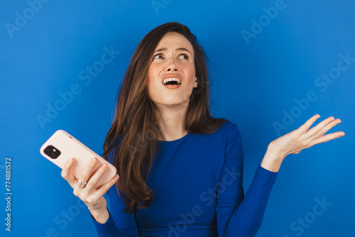 Mad and shock young brunette beautiful woman using mobile phone isolated on blue background. Yelling unhappy woman hold mobile phone. Disappointed sad upset lady horrified impressed news. WTF. Oh no. photo
