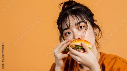Generative AI   Young Asian woman eating hamburger on orange background