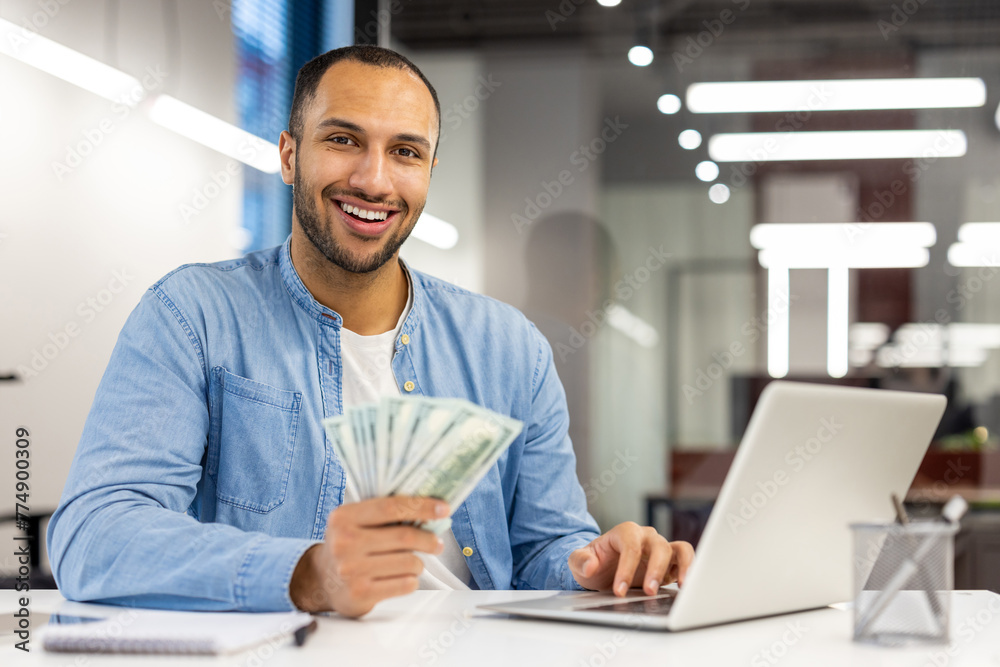 Portrait of a smiling young Latin American man sitting at an office desk, working on a laptop, holding cash banknotes in his hand, looking confidently at the camera Portrait of a smiling young Latin American man sitting at an office desk, working on a laptop, holding cash banknotes in his hand, looking confidently at the camera