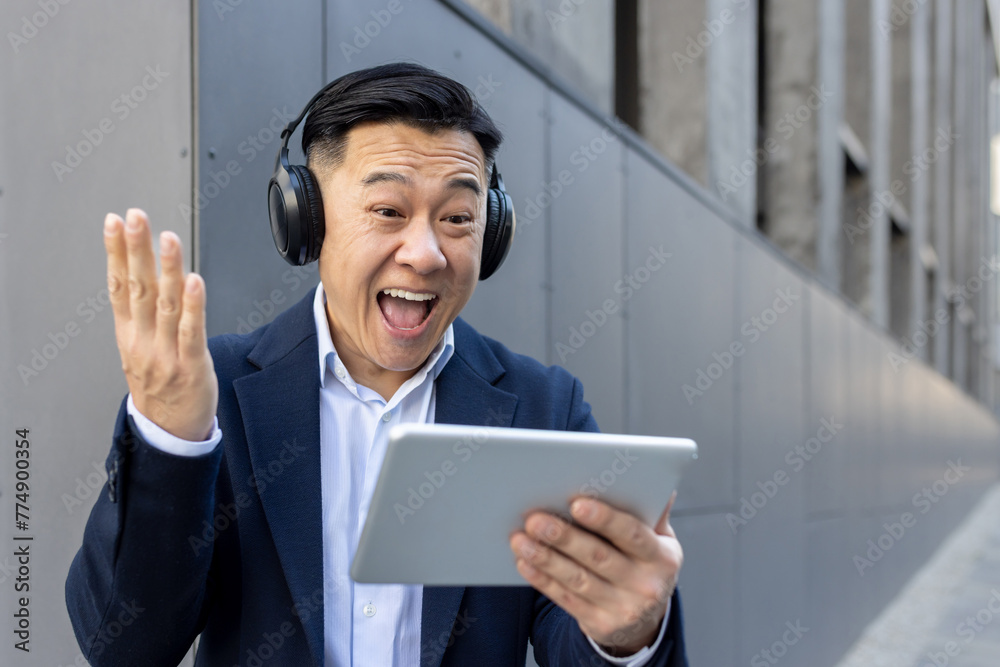 Happy young Asian male businessman standing in headphones outside office center, looking at tablet screen and happy with news and success Happy young Asian male businessman standing in headphones outside office center, looking at tablet screen and happy with news and success