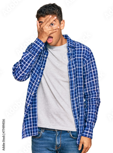 Young handsome african american man wearing casual clothes peeking in shock covering face and eyes with hand, looking through fingers with embarrassed expression.