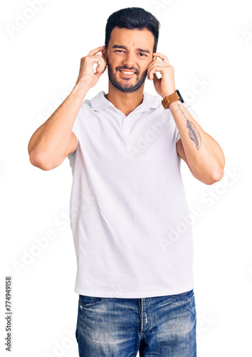 Young handsome hispanic man wearing casual clothes covering ears with fingers with annoyed expression for the noise of loud music. deaf concept.