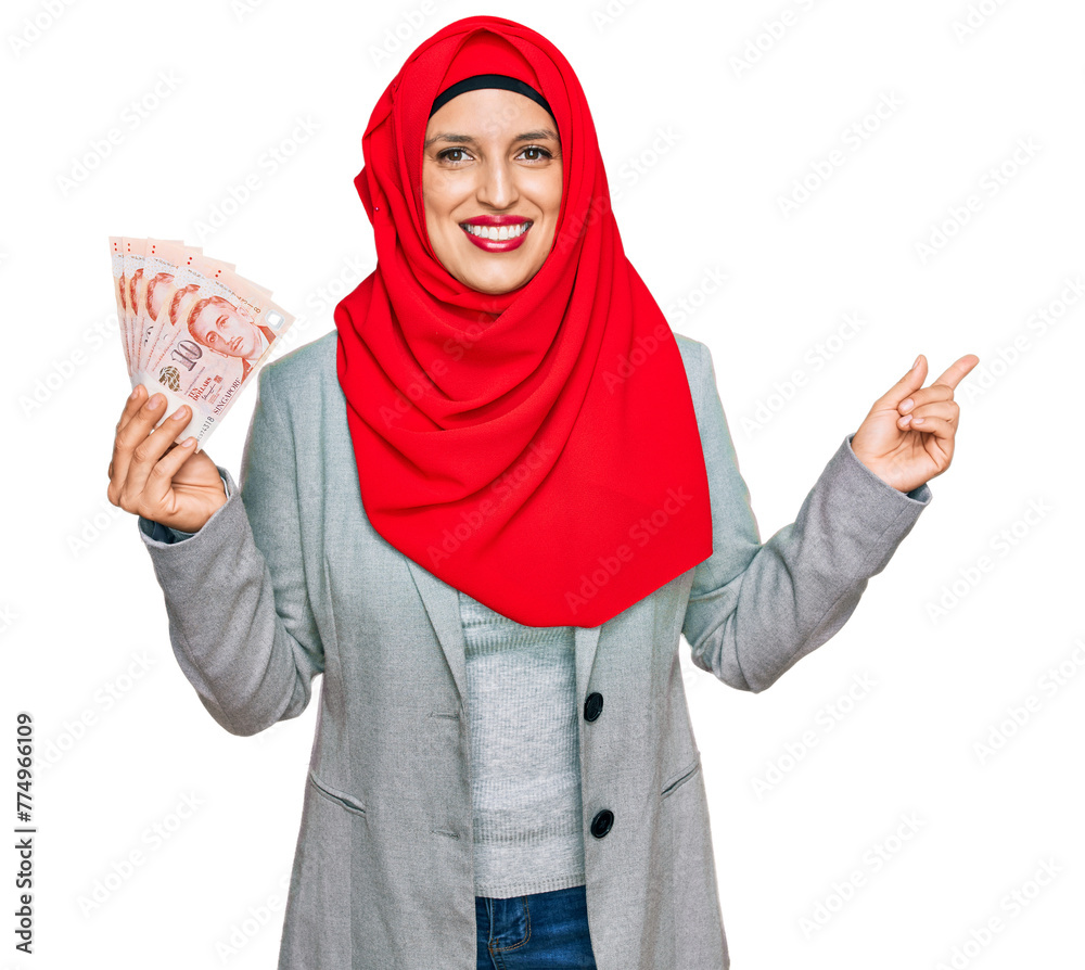 Beautiful hispanic woman wearing islamic hijab holding singapore dollars banknotes smiling happy pointing with hand and finger to the side Beautiful hispanic woman wearing islamic hijab holding singapore dollars banknotes smiling happy pointing with hand and finger to the side