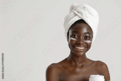 Skincare, cheerful young African American woman smiling, applying moisturizing cream on face, lotion or mask for skin lifting and anti-aging, wears wrapped towel on head, with plastic jar in hand. 