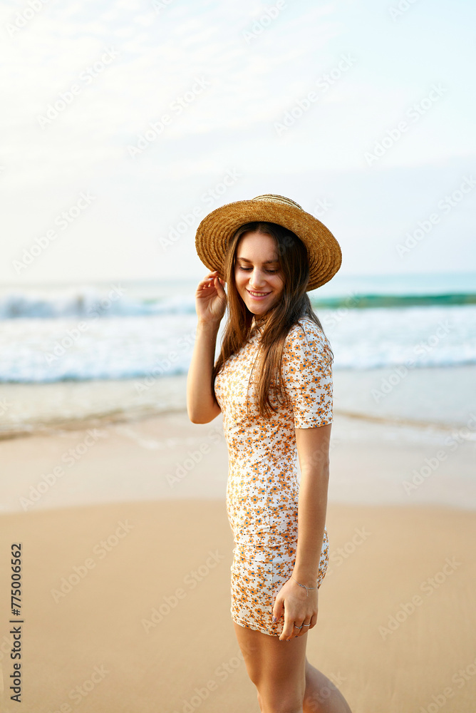 Smiling woman in vintage floral dress and straw hat walks on sandy beach. Blue ocean waves in background. Summer, fashion, leisure concept. Happy female enjoys sunset by the sea, retro style.