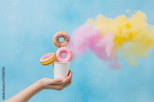 A young woman's hand holds a paper cup into which baby donuts fall from above with colored smoke, light blue background, realistic texture, the donuts are covered with pink and yellow chocolate