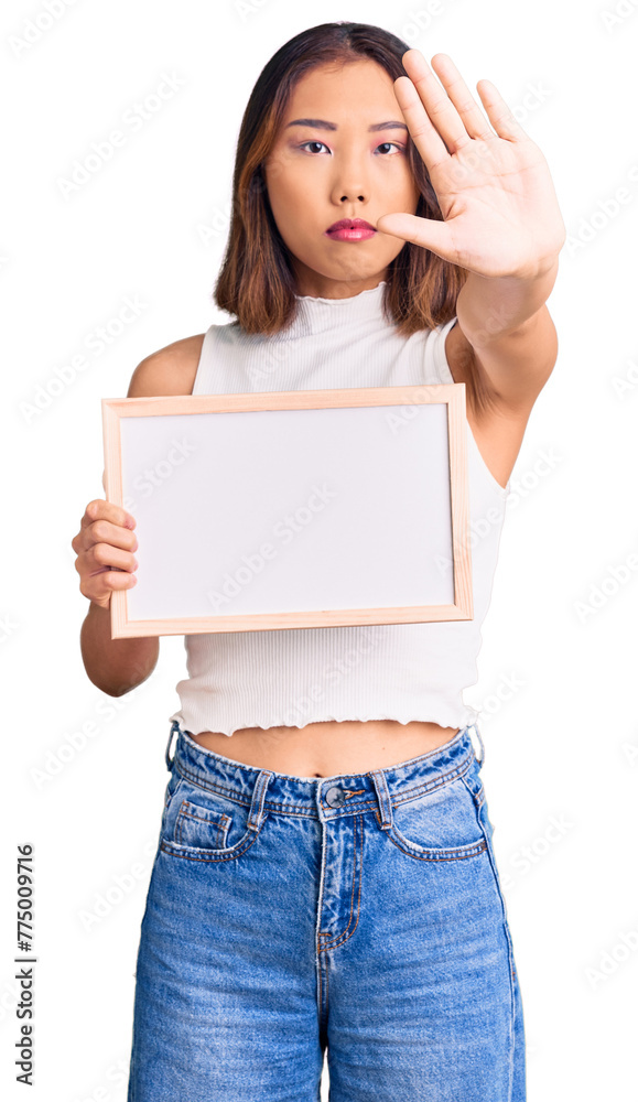 Young beautiful chinese girl holding empty white chalkboard with open hand doing stop sign with serious and confident expression, defense gesture Young beautiful chinese girl holding empty white chalkboard with open hand doing stop sign with serious and confident expression, defense gesture