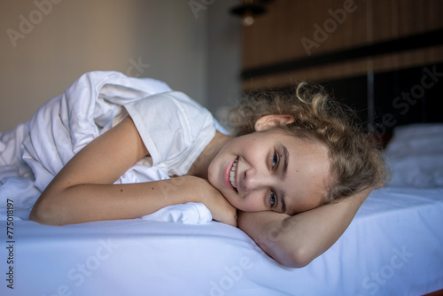 A cute portrait of a blonde teenage girl in a white blanket on a large comfortable bed after a night's rest in a beautiful cozy hotel room.