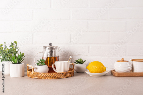 Beautiful kitchen background with glass teapot of fragrant tea, cup, lemon on wicker tray. Front view. White brick wall. A copy space.