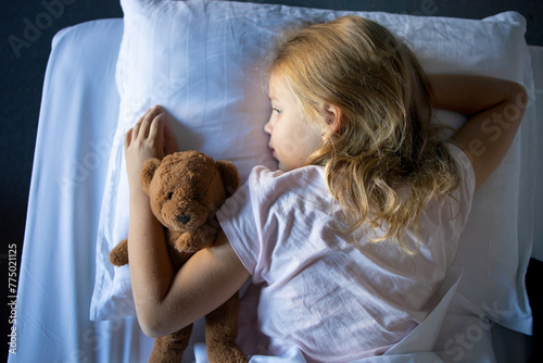 An overhead view of a sleeping Little blonde girl in pajamas on a large and cozy bed, she hugs her toy teddy bear. Rest and proper sleep in children