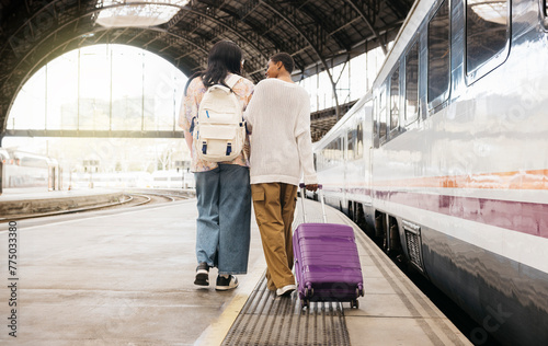 Rear view of two young black women pulling a suitcase and walking together on the platform of a train station to catch a train for a trip.