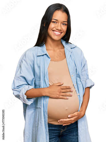 Beautiful hispanic woman expecting a baby showing pregnant belly looking positive and happy standing and smiling with a confident smile showing teeth
