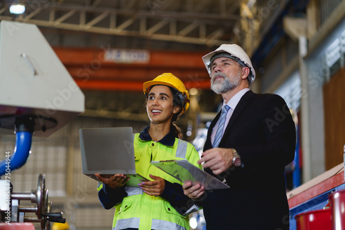 A man and a woman are standing in a factory, looking at a laptop. They seem to be discussing something important about machine in factory control.