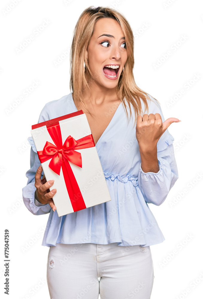 Beautiful blonde woman holding gift pointing thumb up to the side smiling happy with open mouth Beautiful blonde woman holding gift pointing thumb up to the side smiling happy with open mouth