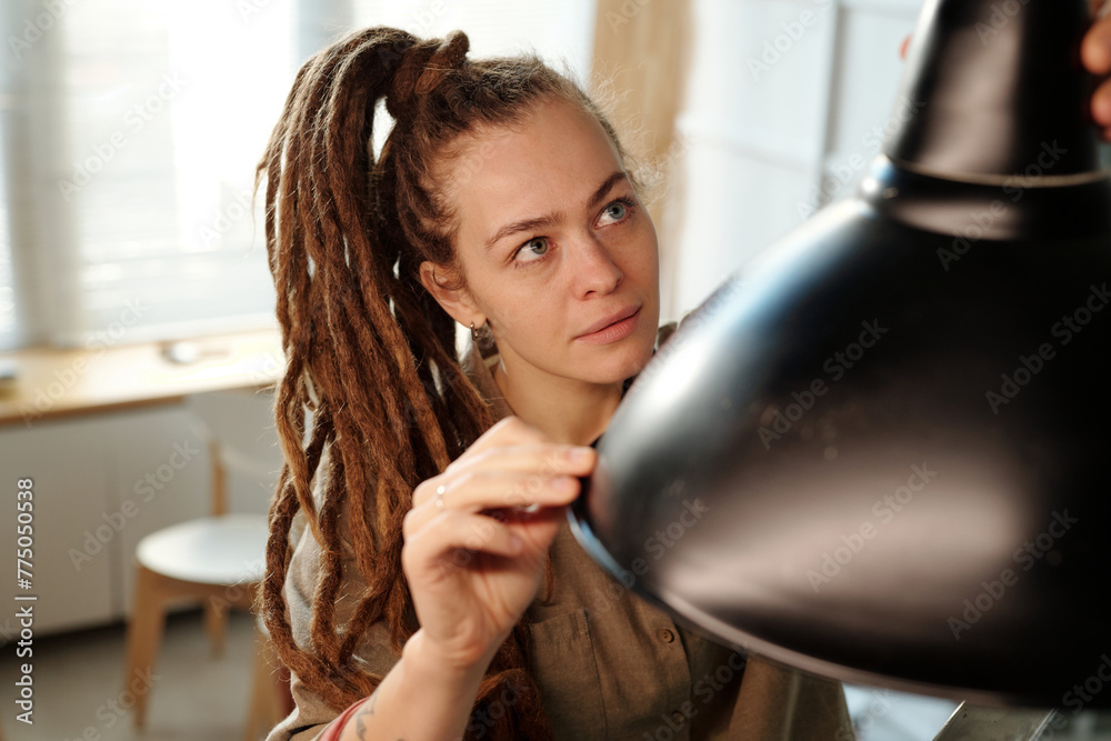 Young female owner of cafe or restaurant with dreadlocks looking at new lamp while hanging it over table during renovation works