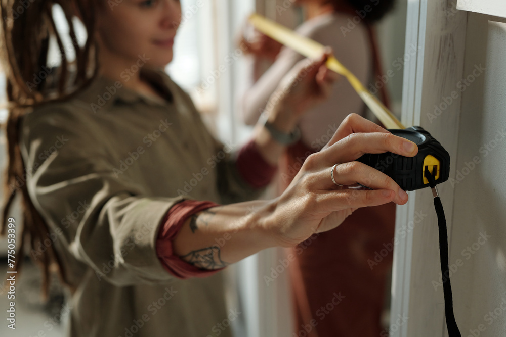 Hand of young female worker or designer of interior taking measures of doorway with measuring tape while standing against colleague