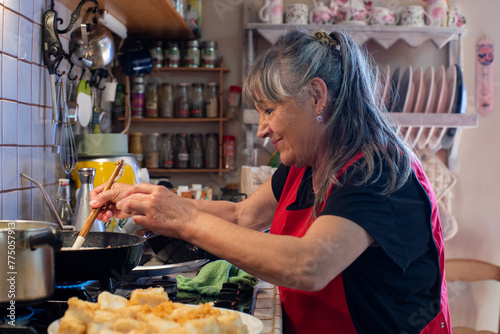 Photos Woman cooking in rustic kitchen, retired person in her sixties with long grey hair, wearing red apron, smiling and stirring with wooden spoon in frying pan