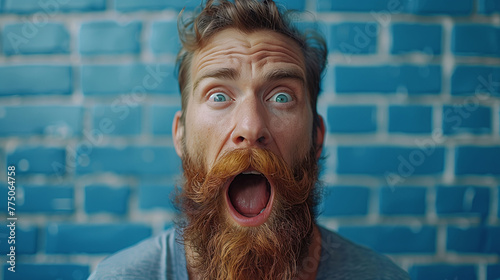 Shocked bearded man with wide eyes and open mouth against a blue brick wall background.