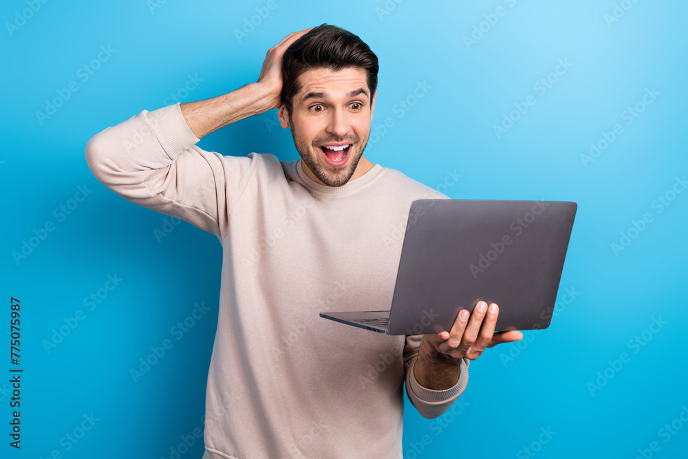 Portrait of young impressed man use laptop unbelievable reaction wear beige sweatshirt isolated on blue color background Portrait of young impressed man use laptop unbelievable reaction wear beige sweatshirt isolated on blue color background