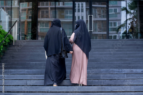 Muslim women in Nigab traditional clothing walks around the city, walking up the stairs into a shopping mall, view from behind