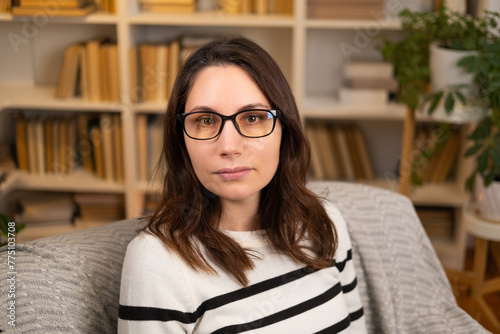 Portrait of a young adult beautiful woman in glasses and a striped blouse sitting in an sofa and looking at the camera indoors