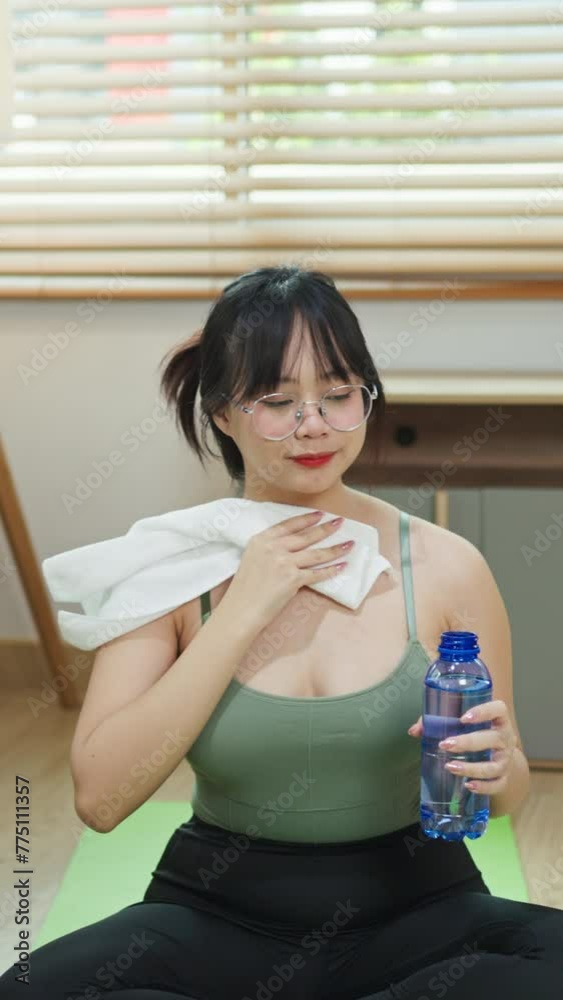 Chubby young woman wiping sweat and drinking water during morning exercise at home Chubby young woman wiping sweat and drinking water during morning exercise at home