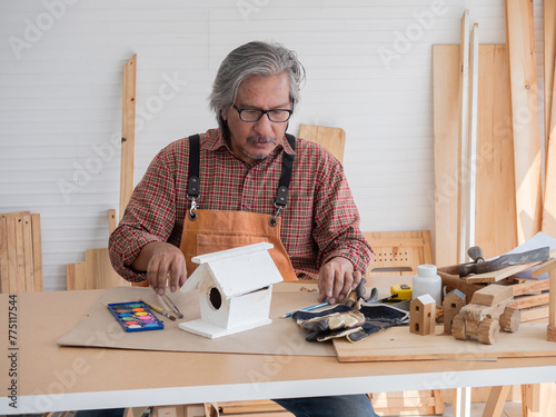 Happy Asian senior man is enjoying his hobby by diligently painting birdhouses in the workshop.