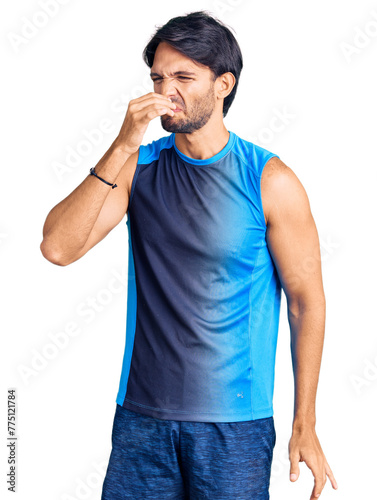 Handsome hispanic man wearing sportswear smelling something stinky and disgusting, intolerable smell, holding breath with fingers on nose. bad smell