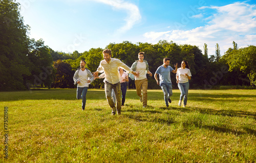 Group of happy friends having fun in park on summer holidays. Several young people spending time in nature. Cheerful, joyful, excited men and women enjoying freedom and running on green grass in sun #775121988