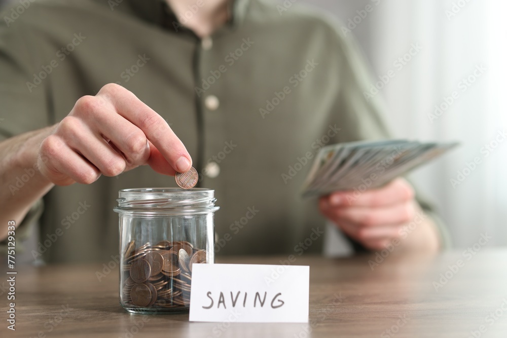 Fototapeta premium Financial savings. Man putting coin into glass jar at wooden table, closeup