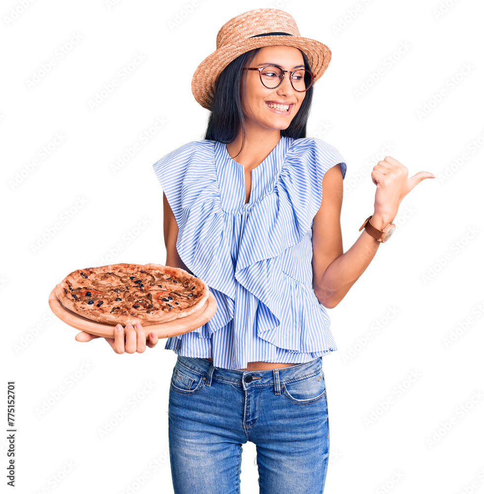 Fototapeta premium Young beautiful latin girl wearing summer hat and glasses holding pizza pointing thumb up to the side smiling happy with open mouth