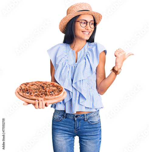 Young beautiful latin girl wearing summer hat and glasses holding pizza pointing thumb up to the side smiling happy with open mouth © Krakenimages.com