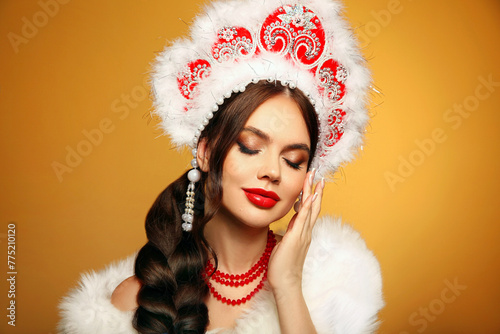 Beautiful brunette fashion woman portrait in fur coat and kokoshnik (woman's headdress in old Russia). Pretty model girl with red lips and plait hairstyle isolated on yellow studio background. photo