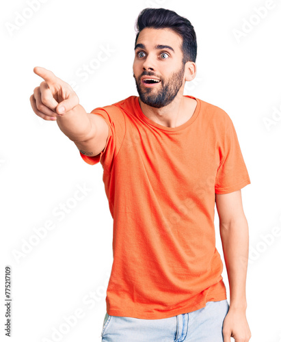 Young handsome man with beard wearing casual t-shirt pointing with finger surprised ahead, open mouth amazed expression, something on the front