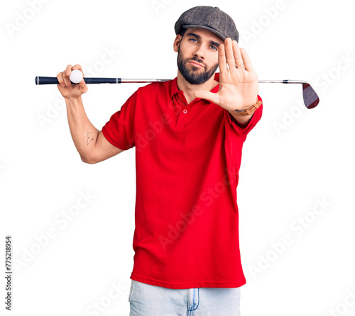 Young handsome man with beard playing golf holding club and ball with open hand doing stop sign with serious and confident expression, defense gesture