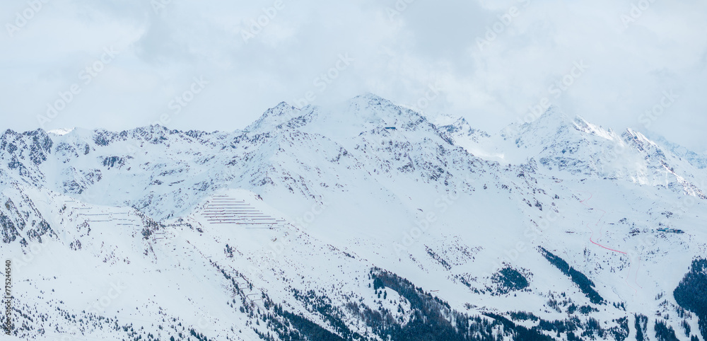 An aerial shot shows Verbier, Switzerland's snowy landscape, ski tracks, a peak, and the Alps under a partly cloudy sky. Its winter charm is in the untouched snow and gentle cloud shadows.