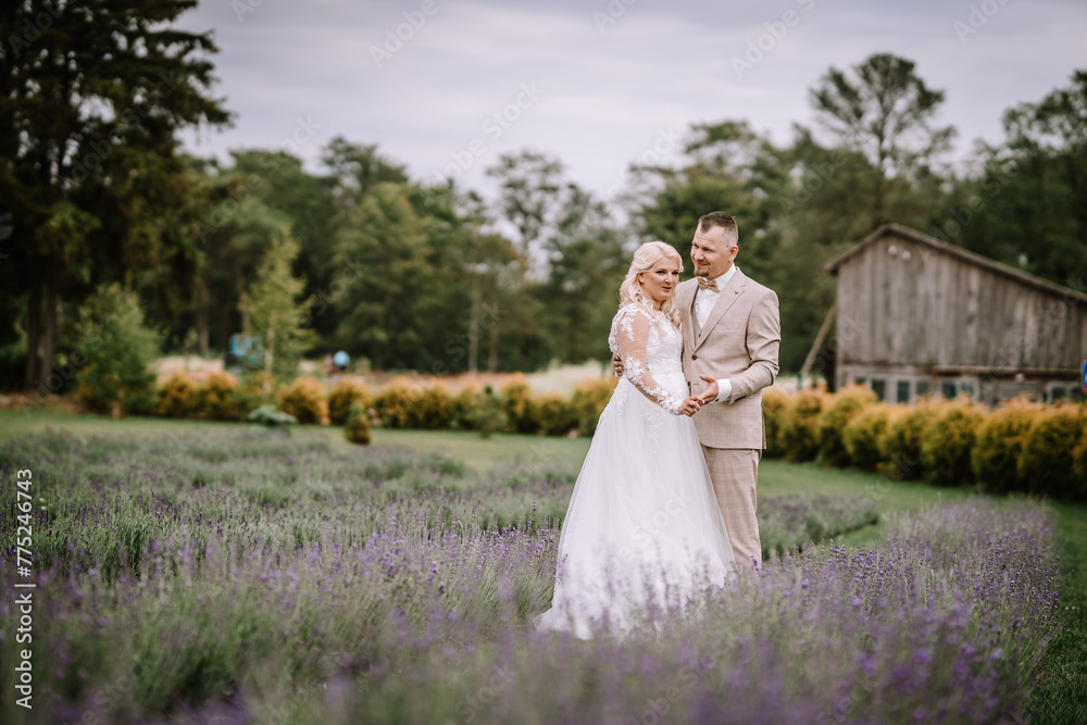 Valmiera, Latvia - Augist 13, 2023 - A bride and groom stand closely, holding hands in a field of lavender, exchanging smiles, with a rustic building and trees in the background. Valmiera, Latvia - Augist 13, 2023 - A bride and groom stand closely, holding hands in a field of lavender, exchanging smiles, with a rustic building and trees in the background.