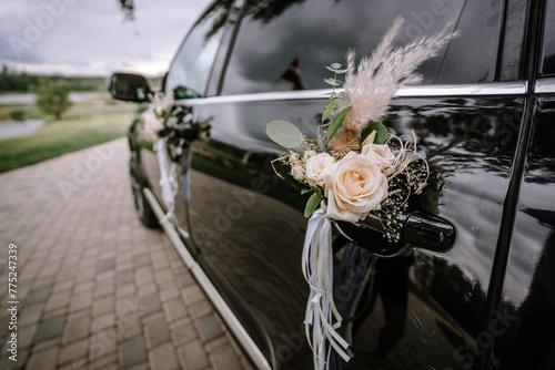 Obraz na plátně A floral decoration with white roses and feathers is attached to the door handle of a black car, likely part of a wedding convoy