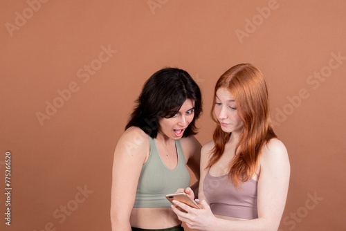 Two girls in sportswear gaze at a phone against a brown backdrop. One wears a surprised expression, hinting at an unexpected revelation, while the other watches with curiosity.