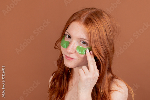 Red-haired woman with under-eye treatment patches on a brown background, looking at camera.