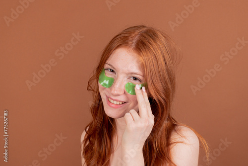 Smiling young woman with red hair applying green under-eye patches on a beige background.
