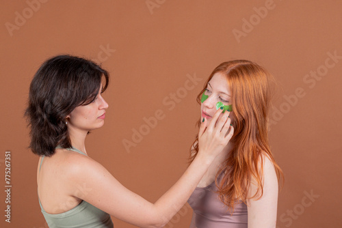 A brunette girl applies green eye patches to a red-haired model, both dressed in sportswear, against a brown background.