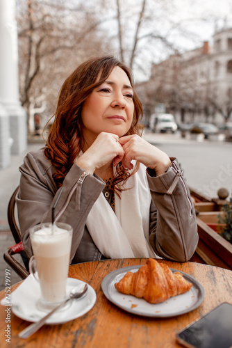 A charmig middle-aged woman drinks latte and eats  croissant in a french cafe on a city street. A woman with flowers is enjoing a wonderful and tasty breakfast photo