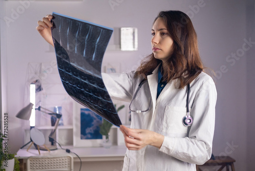 A young adult white female doctor in a medical mask and a white coat holds a CT scan in her hands and looks at it carefully