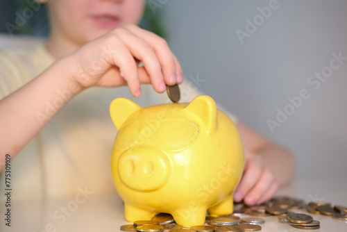 Child's hand folded money coin into piggy bank. Pile of coins, business, finance and saving money for future concept. Adorable boy with yellow piggy bank at home. Children's pocket money.