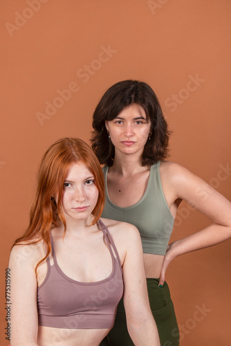 Two young women posing together against a brown background, looking at the camera with a confident expression.