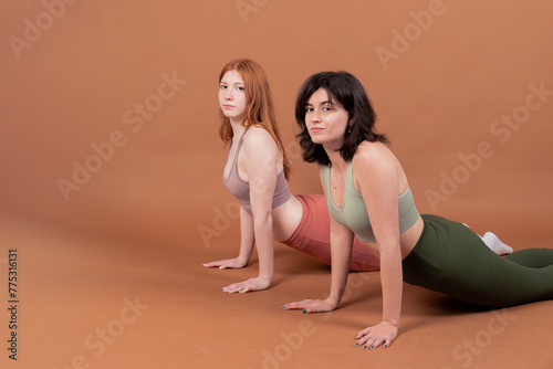 Two young girls dressed in yoga sportswear perform the Cobra Pose against a brown background, showcasing strength and flexibility in their practice.